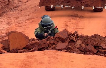 Persona con traje de protección y casco, agachada sobre rocas en un entorno de tierra roja, examinando un dispositivo explosivo.