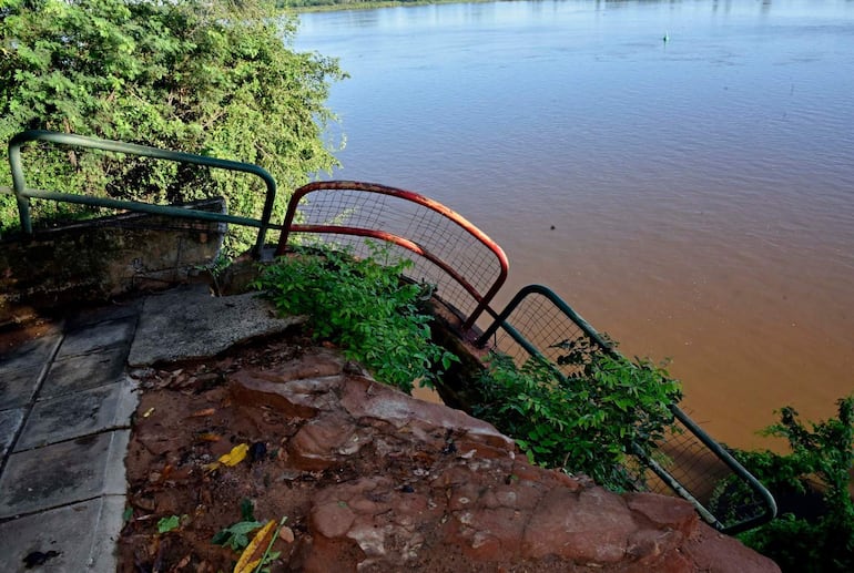 La imagen muestra un entorno natural, específicamente un área adyacente a un río. En la escena no hay personas visibles. Como elemento central, se observa una baranda de metal, parte de la cual es de color verde y otra parte está pintada de rojo, y parece deteriorada. A la izquierda se aprecia un camino de baldosas desgastadas que conduce a la orilla del río, visible en el fondo. La vegetación alrededor es densa, con arbustos y plantas que crecen cerca del agua. No se identifican símbolos de algún equipo de fútbol ni textos visibles que aporten contexto adicional. El ambiente es exterior y parece ser un área de acceso al río, posiblemente un mirador o un parque, aunque no hay monumentos o estructuras icónicas en la imagen. La escena sugiere tranquilidad, con el agua calmada y un clima despejado.