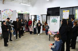 Mujer con gafas sostiene un documento mientras habla, rodeada de adultos y niños en un ambiente festivo decorado con carteles.