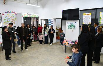 Mujer con gafas sostiene un documento mientras habla, rodeada de adultos y niños en un ambiente festivo decorado con carteles.