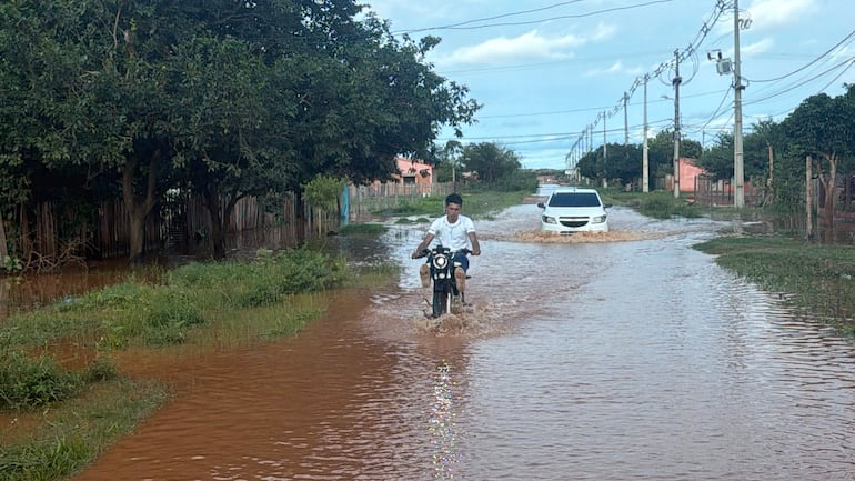 Un joven en motocicleta con camisa blanca y pantalones oscuros circula por una calle inundada, levantando agua.