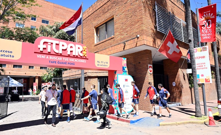 Siete hombres jóvenes en camisetas rojas y azules, caminando hacia el evento FITPAR 2023, con edificio de ladrillos y banderas de fondo.