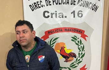 Hombre de pie en chaqueta oscura y camiseta verde, junto a banner de Policía Nacional en una comisaría.