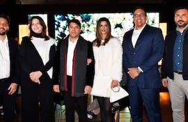 Seis personas conversan en un ambiente elegante; hombres y mujeres con trajes formales y abrigos, sonriendo en un interior moderno.