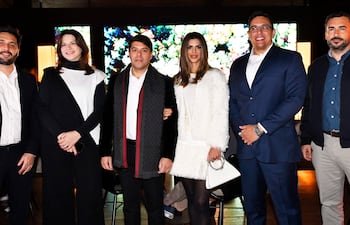 Seis personas conversan en un ambiente elegante; hombres y mujeres con trajes formales y abrigos, sonriendo en un interior moderno.