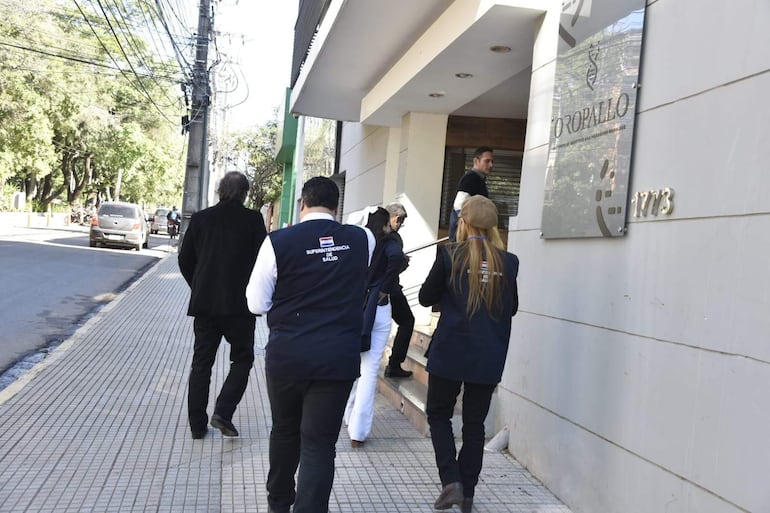 Grupo de personas, dos de espaldas: uno con traje negro y otro con chaleco de la Superintendencia de Salud, caminan hacia un edificio.