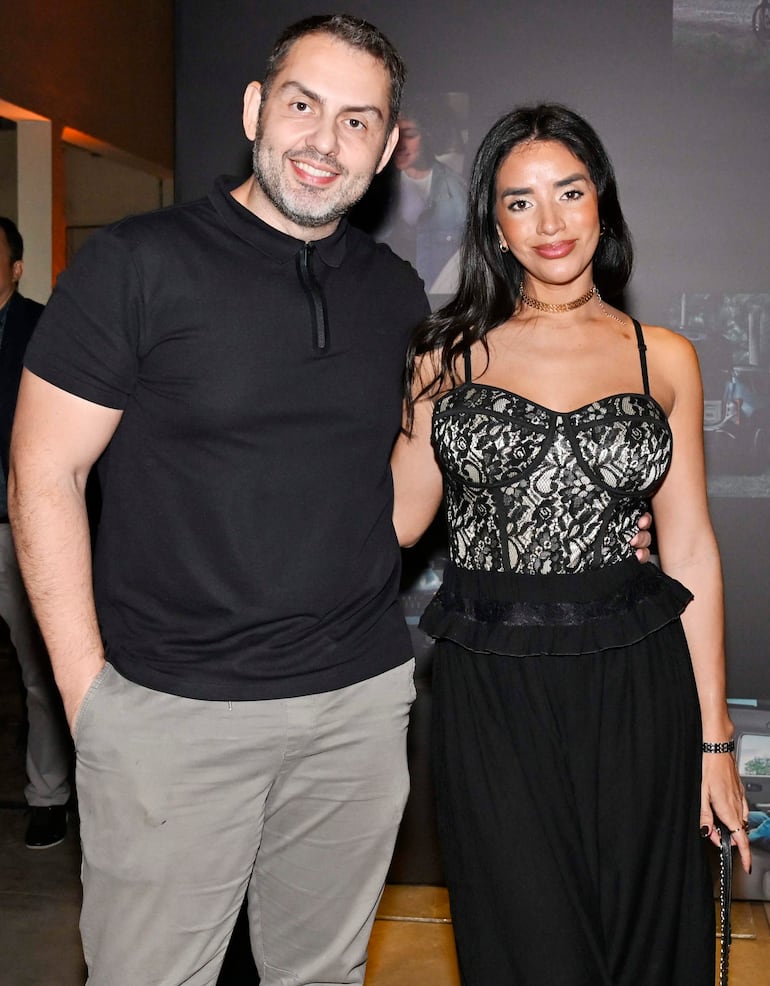 Hombre con camiseta negra y pantalones claros, y mujer en vestido negro con encaje, sonriendo en un evento moderno.