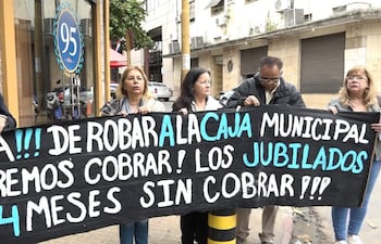 Cinco manifestantes, mujeres y hombre, sosteniendo carteles en protesta con expresiones de tristeza frente a un edificio.