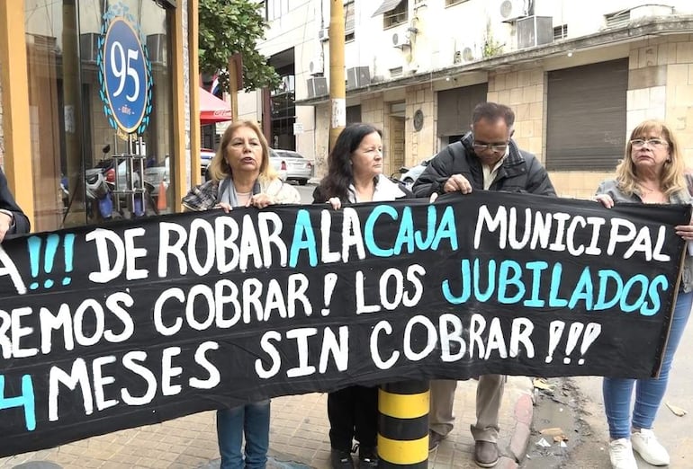 Cinco manifestantes, mujeres y hombre, sosteniendo carteles en protesta con expresiones de tristeza frente a un edificio.