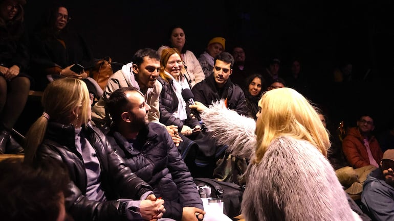 Mujer con abrigo de piel artificial sostiene micrófono mientras interactúa con el público, que observa atentamente en ambiente oscuro.
