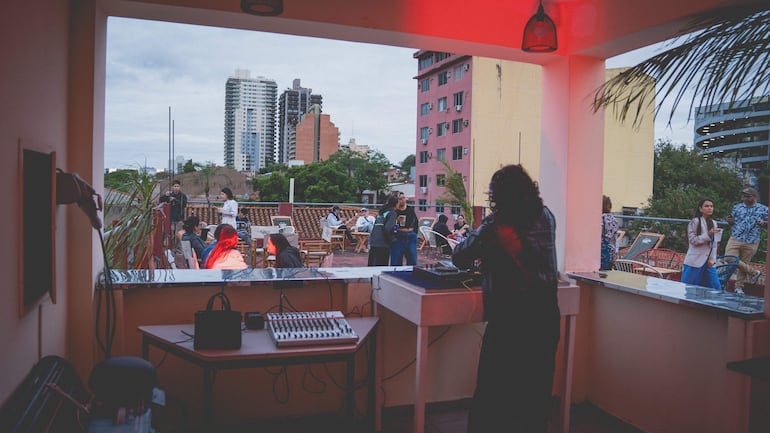 Grupo de personas en terraza. Una persona de espaldas manipula equipo de sonido, otros conversan y disfrutan del ambiente urbano.