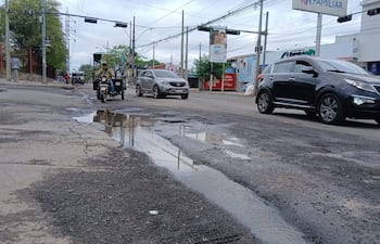 Avenida Artigas con baches y charcos, motociclista a la izquierda, coches en movimiento, cielo nublado y ambiente urbano evidente.