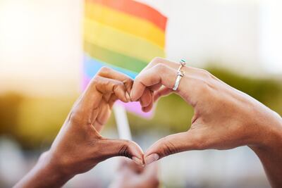 Dos mujeres forman un corazón con las manos y detrás flamea una bandera del orgullo gay con los colores del arcoiris.
