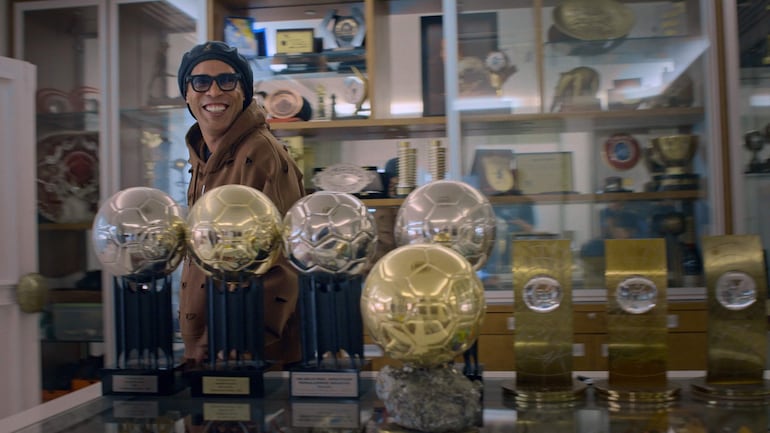 Ronaldinho con chaqueta marrón y gorro negro sonríe ante la cámara, rodeada de trofeos, incluidos tres balones dorados.