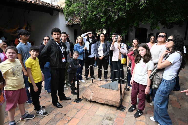 Un grupo diverso de personas, incluyendo niños, observa con atención una piedra mientras una mujer graba con su móvil.