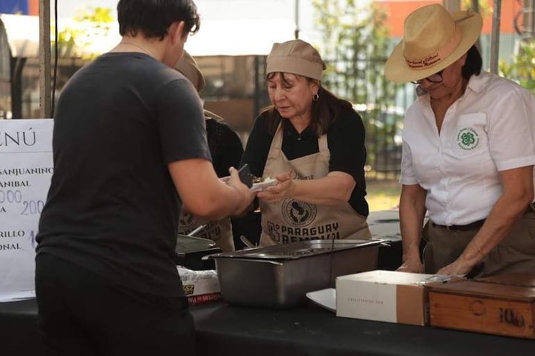 Dos mujeres en chaquetas negras y blancas, con sombreros, sirven alimentos a un hombre joven en un ambiente al aire libre.