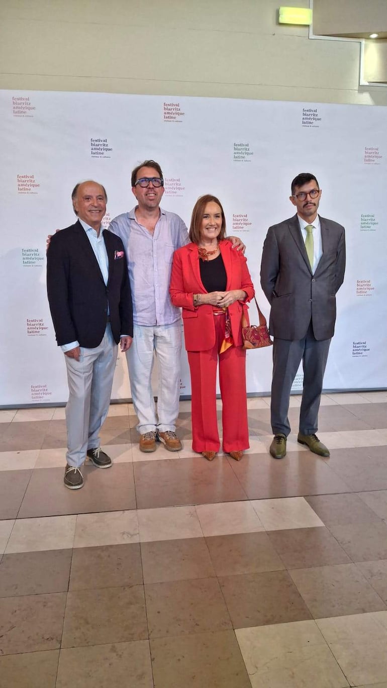 Cuatro personas posan sonrientes. Un hombre vestido con un blazer oscuro y pantalón claro, una mujer en conjunto rojo y dos hombres más en traje, todos en un salón.