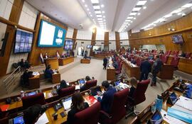 Varios legisladores en el recinto legislativo, sentados en bancos rojos, con laptops y documentos en mesas.