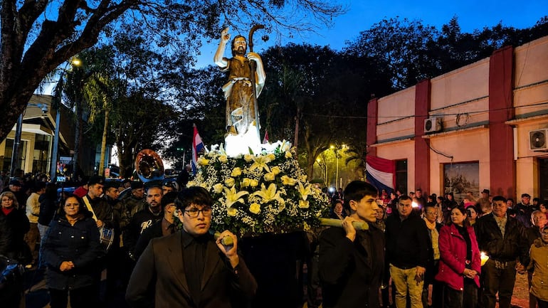 Jóvenes en trajes oscuros sostienen la base de una estatua, mientras el público observa con respeto en un ambiente iluminado con antorchas.