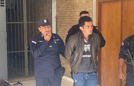 Juan Antonio González Ferreira con abrigo oscuro y jeans, mientras es observado por un oficial de policía en uniforme azul.