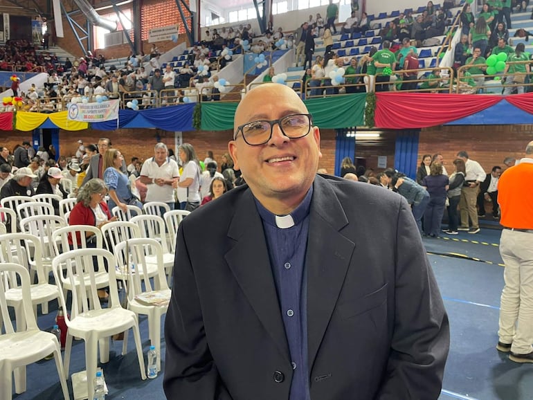 Hombre con gafas y traje oscuro conversa en evento festivo, rodeado de público en un gimnasio decorado con globos y banderas.