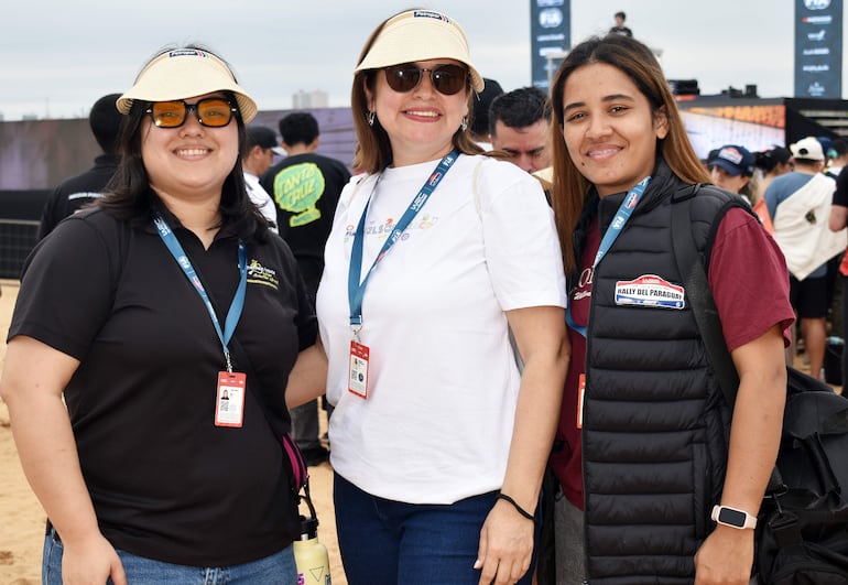 Tres mujeres sonríen juntas, una con camiseta negra, otra con blanca y la última con chaqueta negra, en un entorno de evento al aire libre.