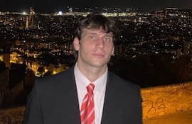 Gaspi en traje negro y corbata roja, de pie con fondo urbano iluminado y expresión tranquila.