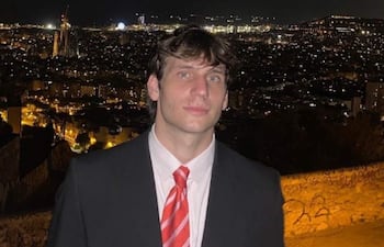 Gaspi en traje negro y corbata roja, de pie con fondo urbano iluminado y expresión tranquila.