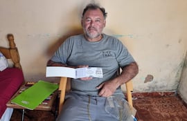 Hombre sentado en silla de mimbre con camiseta gris Nike, sosteniendo un documento, conectado a oxígeno, en habitación beige.