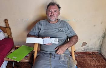 Hombre sentado en silla de mimbre con camiseta gris Nike, sosteniendo un documento, conectado a oxígeno, en habitación beige.