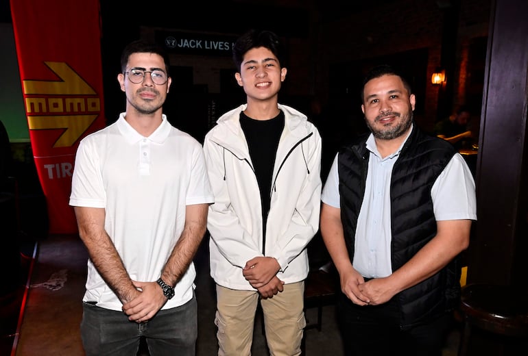 Tres hombres sonrientes: uno con gafas y camiseta blanca, otro con chaqueta blanca y otro en camisa azul y chaleco negro.
