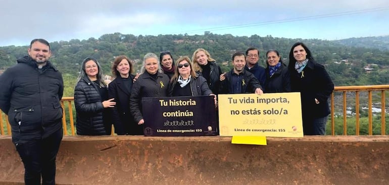 Grupo de personas vestidas de negro con lazos amarillos sostiene carteles que dicen 'Tu vida importa, no estás solo/a' en un ambiente verde.