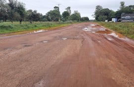Carretera de tierra deteriorada, paisaje gris y nublado, sin personas visibles, rodeada de vegetación. Cartel publicitario a la derecha.