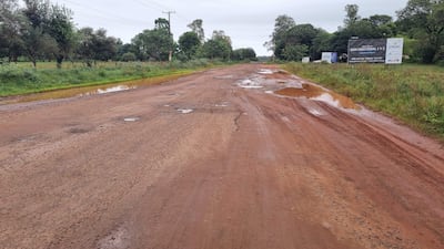 Carretera de tierra deteriorada, paisaje gris y nublado, sin personas visibles, rodeada de vegetación. Cartel publicitario a la derecha.