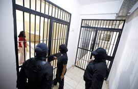 Tres personas de pie frente a una celda observan a una mujer en camiseta roja y pantalones de cuadros, dentro de la prisión.