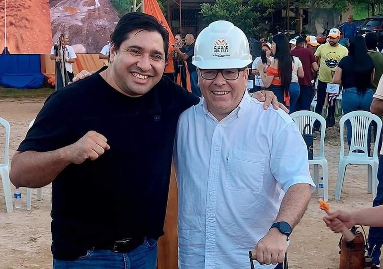 Hombre con casco de obra sonríe junto a otro de camiseta negra, ambos en un evento al aire libre con multitud de fondo.