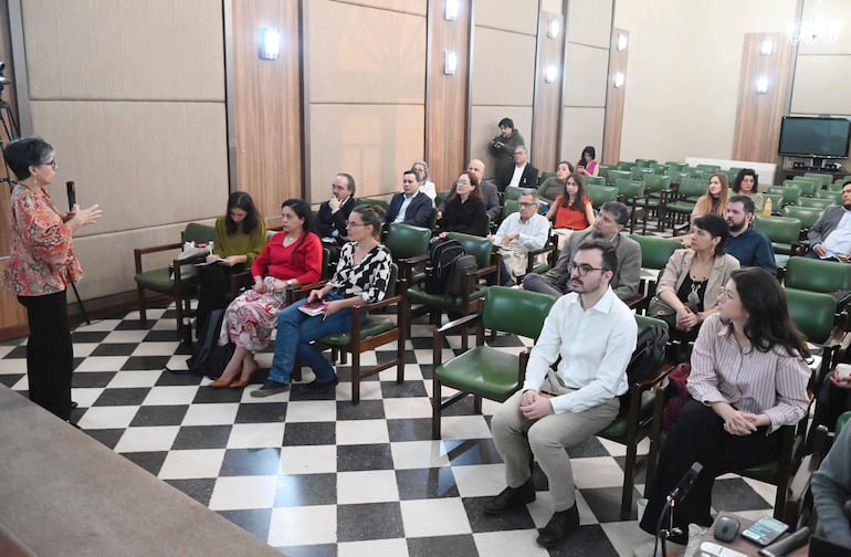Oradora en blusa de colores cálidos gesticula frente a audiencia atenta en salón con piso a rayas.