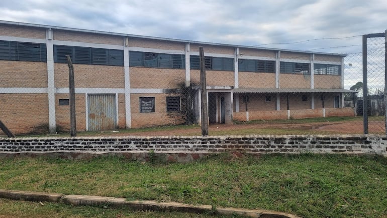 Edificio de ladrillos en estado de abandono, rodeado de cerca de alambre y área ajardinada, con cielo nublado.