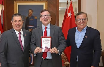 Horacio Cartes en traje gris oscuro sostiene su libro, mientras José Duarte Penayo y Oscar Nicanor Duarte Frutos sonríen en un despacho con banderas.