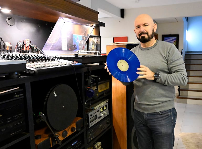 Jorge Puig con suéter gris y jeans, sosteniendo un disco de vinilo azul frente a un equipo de sonido en un ambiente iluminado.