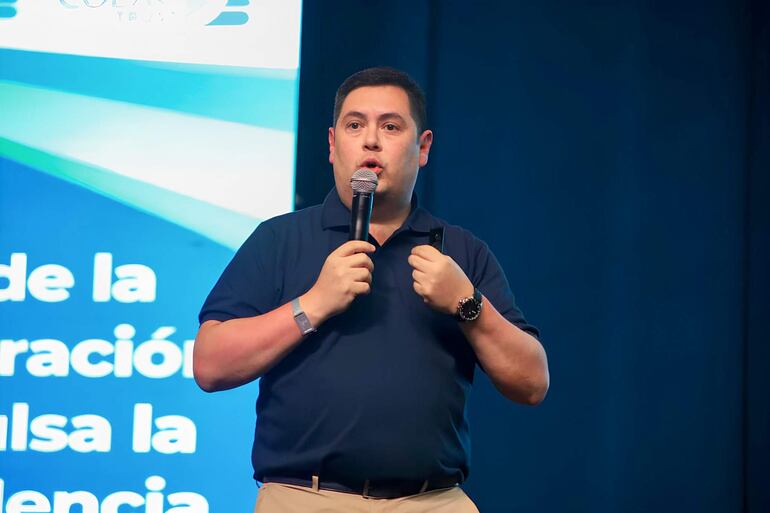 Hombre en camiseta oscura sostiene micrófono en una conferencia con fondo gráfico que incluye texto parcial.