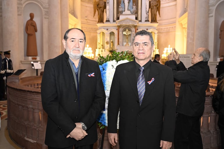 Dos hombres serios en trajes formales frente a un altar con ofrenda floral y decoraciones en el pecho.