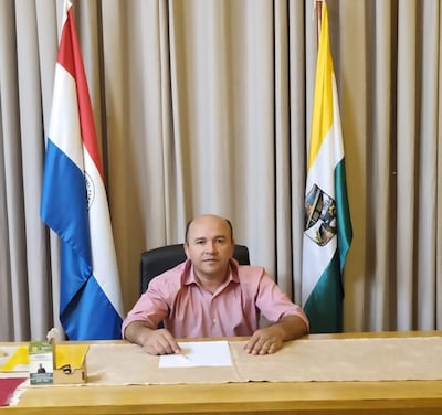 Ramón Sanabria sentado en un escritorio, vistiendo camisa rosa, con banderas de Paraguay y otra detrás.