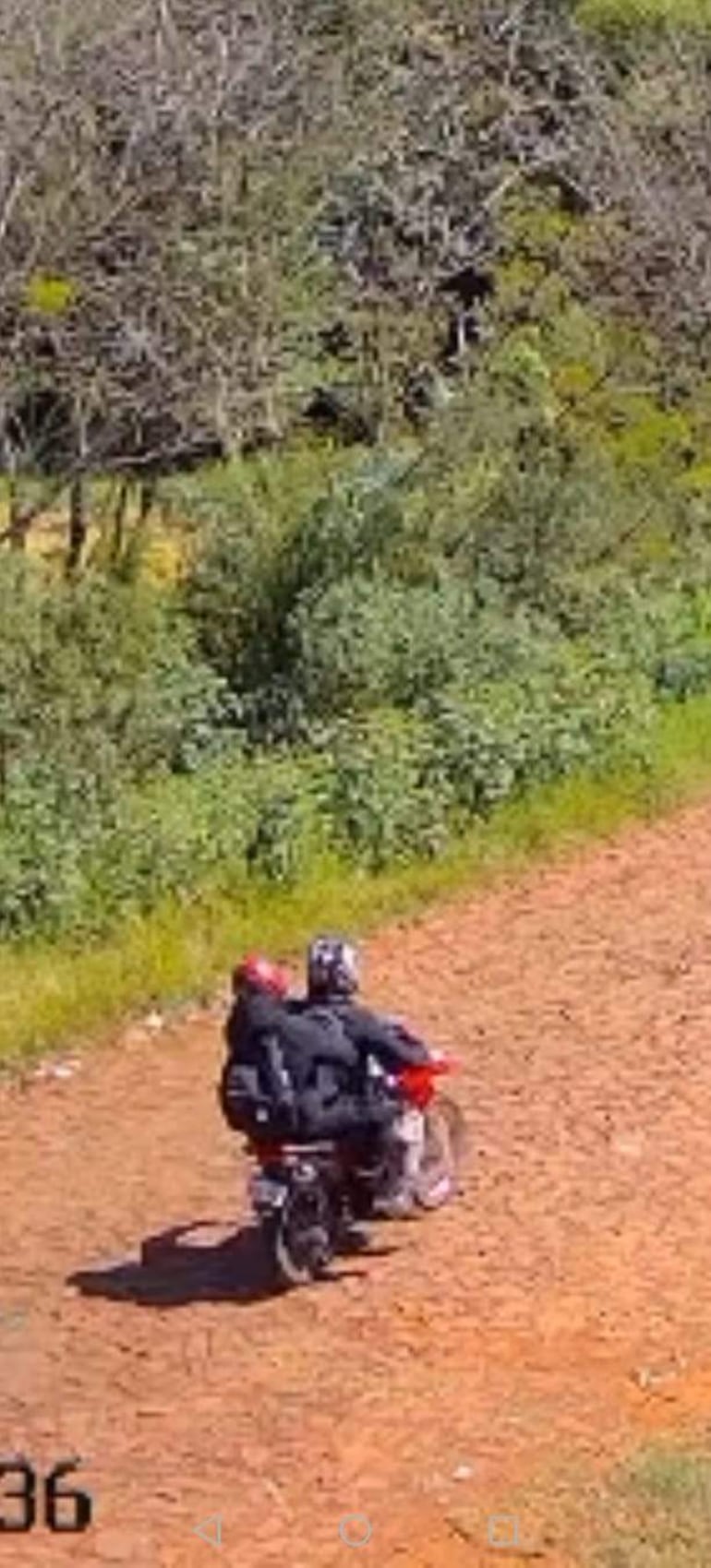 Dos personas con cascos rojo y negro sobre una motocicleta en un camino de tierra rodeado de vegetación.