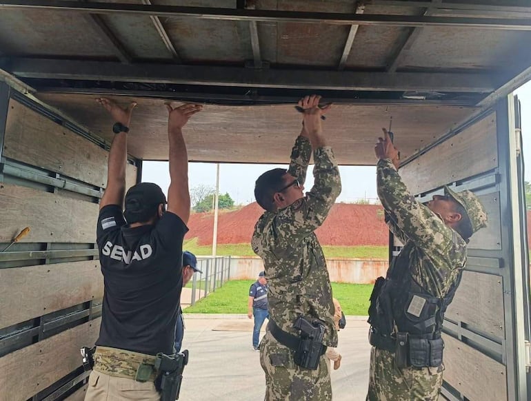Tres personas en camión: dos con uniformes militares y una con camiseta 'SENAD', ajustando equipo en un entorno al aire libre.
