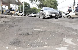 Motociclista con bolso observa la calle deteriorada con baches y vehículos en movimiento, mientras una estación de servicio se asoma al fondo.