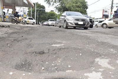 Motociclista con bolso observa la calle deteriorada con baches y vehículos en movimiento, mientras una estación de servicio se asoma al fondo.