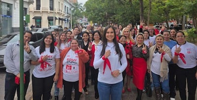 Romina Medina con camiseta blanca y banda roja, rodeada de personas sonrientes en un evento exterior con árboles y edificios de fondo.