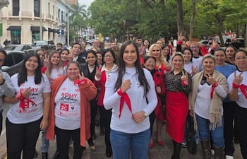 Romina Medina con camiseta blanca y banda roja, rodeada de personas sonrientes en un evento exterior con árboles y edificios de fondo.