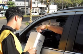 Individuo con chaleco amarillo ofrece una caja a un conductor de coche. Entorno urbano con edificios y vehículos visibles.
